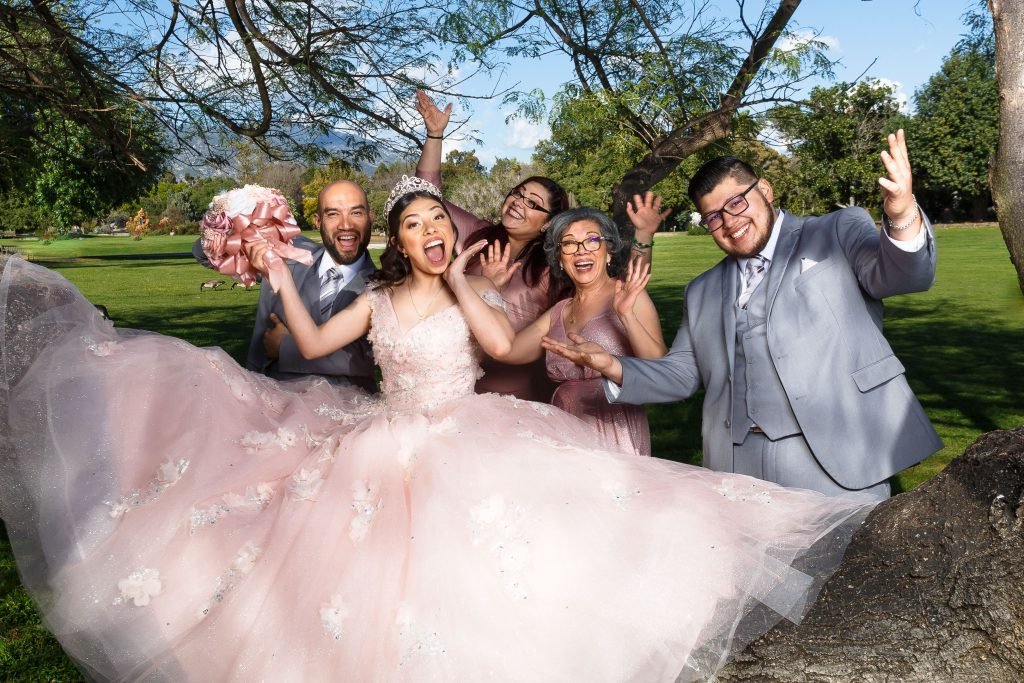 quinceañera en el parque En Los Ángeles, California, bajo un cielo azul y entre árboles frondosos, una quinceañera irradia felicidad junto a su familia, con risas y brazos abiertos, listos para celebrar un día inolvidable.